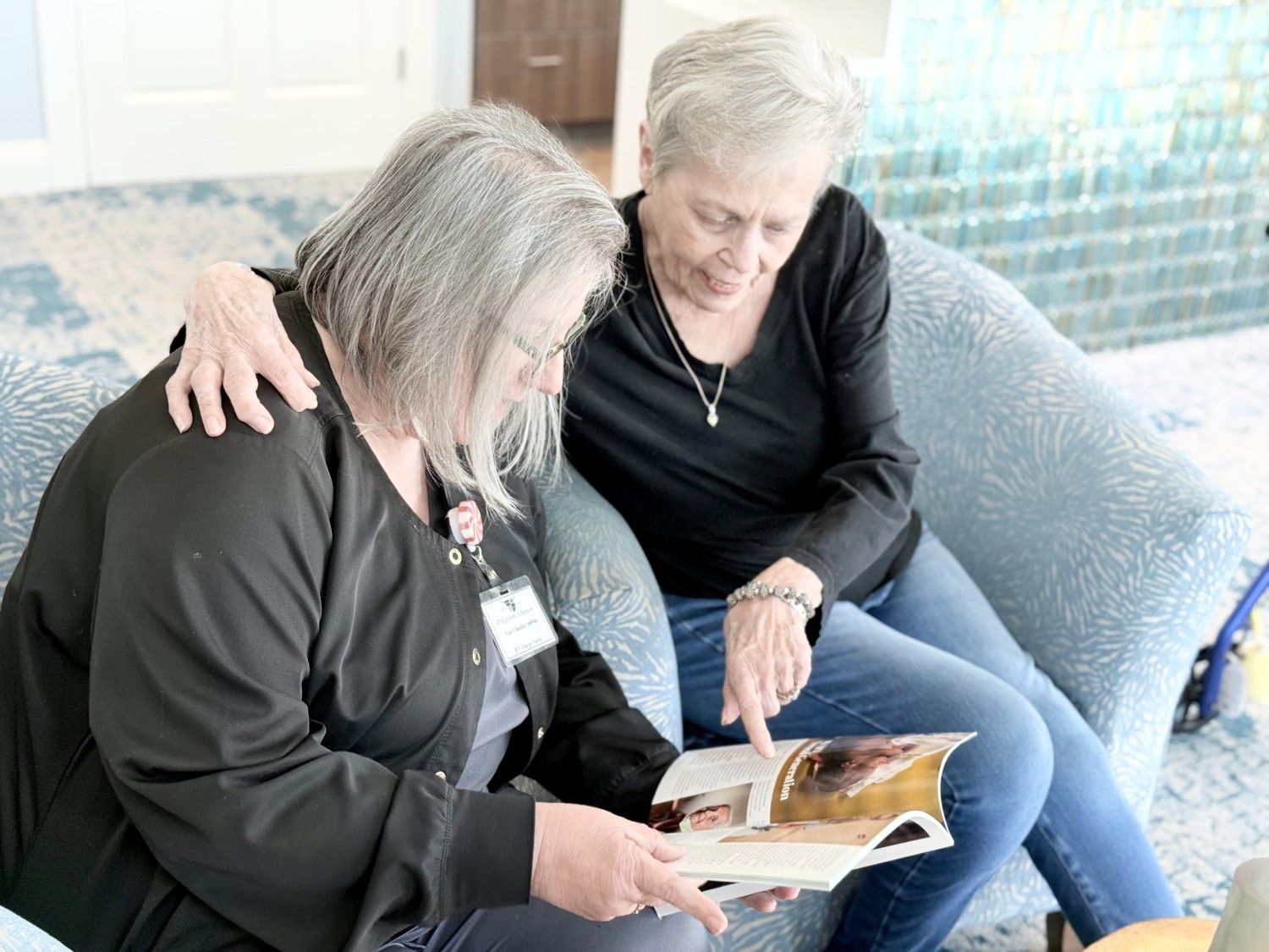 Two women share a magazine on a couch at Pilgrim Manor Skilled Nursing in Bossier City, LA